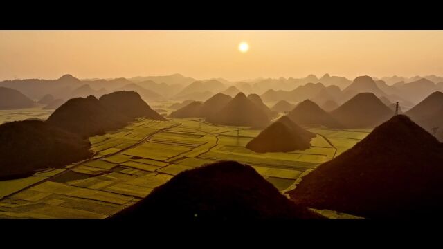 夕阳笼罩金鸡峰丛 黄金铺满螺蛳花田 层峦叠嶂那色峰海 十级泻落九龙瀑布 云雾藏身于鲁布革三峡 竹筏悠扬在多依河上 这里便是古今犹美的多彩罗平